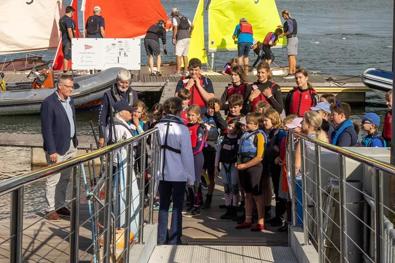 The Princess Royal, seen from behind in a white jacket, talking to a large group of young sailors in buoyancy aids on the Royal Lymington Yacht Club pontoon, with dinghies and helpers behind them.